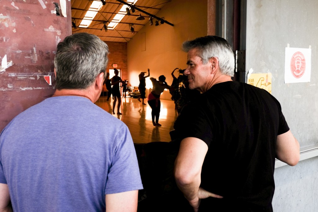 Color photo, two men observing a group of dancers in silhouette.
