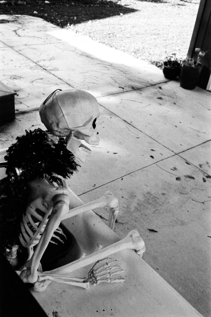 Black and white photo of a human skeleton sitting in a cast iron sink on a patio. Most likely taken around halloween. 